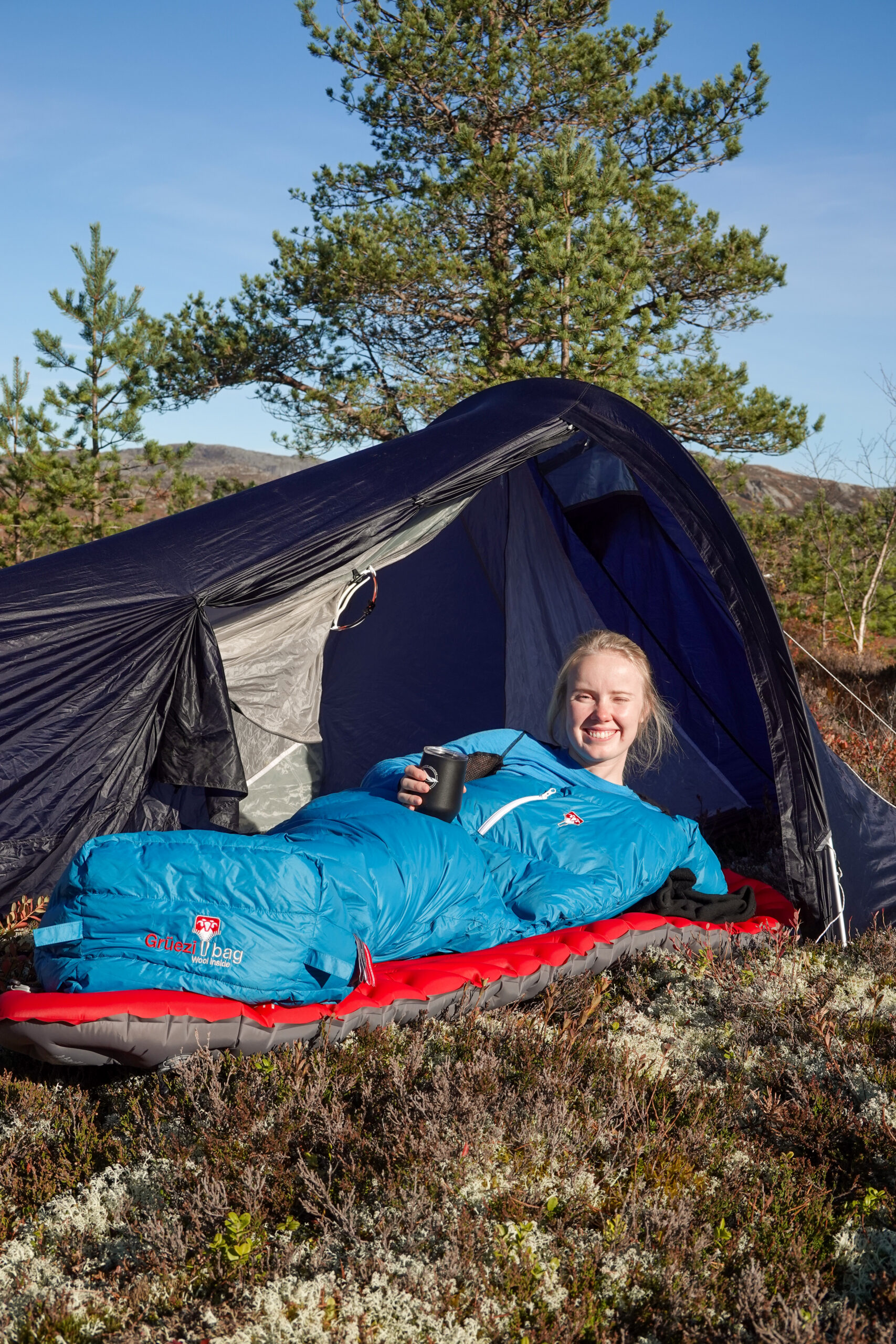 Karen Kyllesø smiler fra en blå Grüezi Bag-sovepose foran teltet med en kopp i hånden.